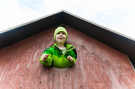 Téléchargez les photos : Un jeune garçon portant un chapeau vert et une veste regarde d'une maison rouge, jouant dans l'aire de jeux. - en image libre de droit