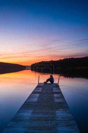 scenic shot of lonely man sitting at wooden pier during sunset