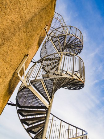 This image captures the intricate design of a metal spiral staircase ascending alongside the exterior of a beige building under a clear blue sky.
