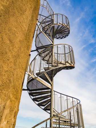 This image captures the intricate design of a metal spiral staircase ascending alongside the exterior of a beige building under a clear blue sky.