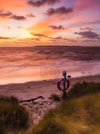 Dieses Bild fängt einen ruhigen Strand während des Sonnenuntergangs ein, mit einem bunten Himmel und einem Rettungsring am Uferrand.