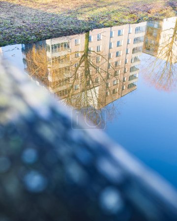 Ein ruhiger Teich in urbaner Umgebung spiegelt ein mehrstöckiges Wohnhaus inmitten der sanften Farbtöne des Morgenlichts wider.
