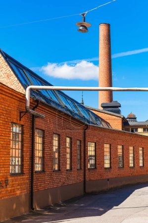 The image showcases an old red brick factory building with a prominent chimney under the clear blue sky, indicating a day of fine weather.
