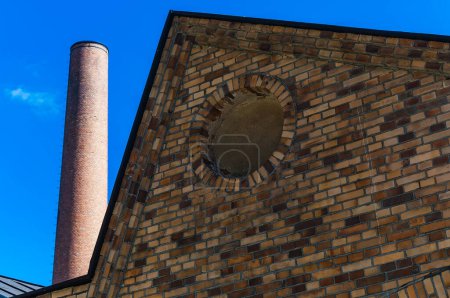 The image captures a segment of a vintage brick building featuring a circular window and a tall, narrow chimney under a bright blue sky.