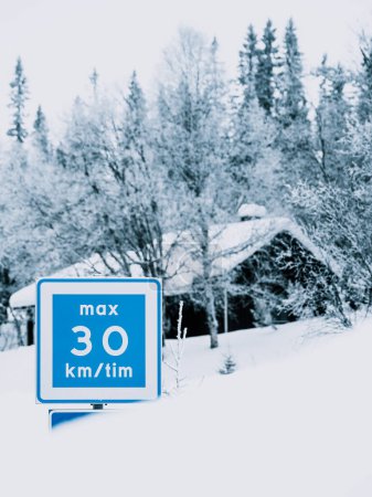 This image captures a tranquil winter scene where a blue speed limit sign displaying max 30 km/tim stands in sharp contrast to the white snow covering the surrounding trees and a partially visible house.