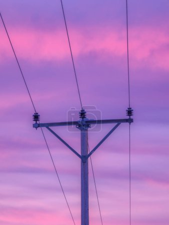 Dieses Bild fängt die Silhouette eines Strommasten ein, der vor einem atemberaubenden Sonnenaufgangshimmel steht, der in Violett- und Rosatönen gemalt ist. Die Stromleitungen sind aus dem Rahmen, was auf ein riesiges Netz der Energieverteilung in der Abenddämmerung hindeutet.