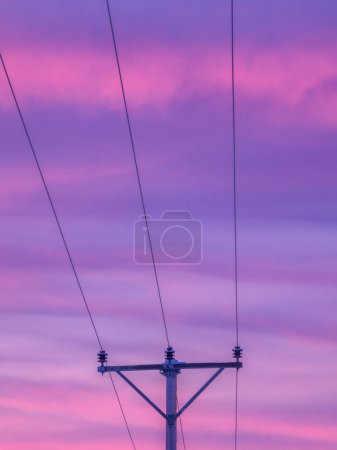 This image captures the silhouette of an electric pylon set against a breathtaking sunrise sky painted in shades of purple and pink. The power lines stretch out of frame, suggesting a vast network of energy distribution at dusk.