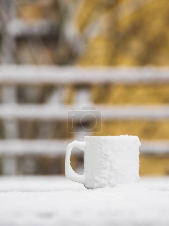In this tranquil scene, a white mug blanketed with fresh snow sits atop a wooden railing, offering a stark contrast to the yellow autumn leaves that adorn the trees softly blurred in the background. The image captures the serene intersection of fall 