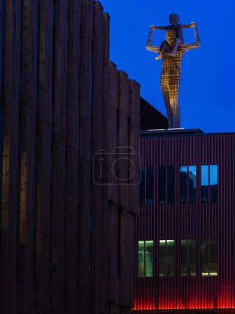 Una llamativa escultura iluminada de una figura humana se alza sobre un edificio moderno con revestimiento vertical de madera. La fotografía captura el contraste entre el cálido resplandor de la escultura y el fresco azul del crepúsculo cielo, así como el architectu