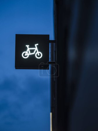 An eye-catching illuminated sign featuring a simple white line drawing of a bicycle stands out against the deep blue hue of the early evening sky. This urban cycling shops signage is designed to attract the attention of passersby and cyclists.