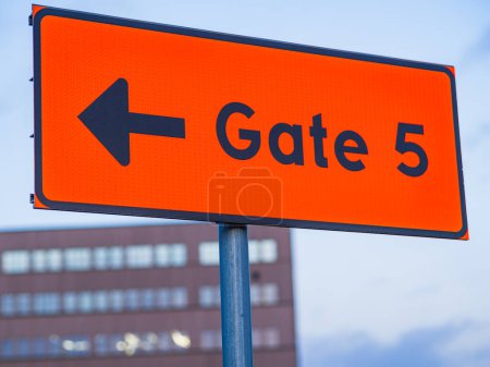 This image captures a vibrant orange sign featuring a arrow pointing left, clearly indicating the direction to Gate 5. The sign stands out against a soft blue sky, with blurred buildings in the background suggesting an urban setting.