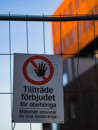 Das Bild zeigt ein schwedisches No-Access-Schild, das an einem Metallzaun befestigt ist und das Verbotssymbol und den Text prominent zeigt, mit einem verschwommenen Gebäudehintergrund während des Abends..