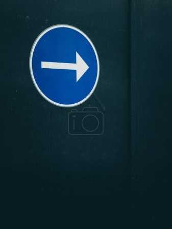 A prominent blue circular sign with a white arrow pointing to the right is centered in the image, affixed to a black surface that fills the background. The sign indicates directional guidance, typically used for traffic or navigation purposes.