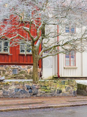 Ein kahler Baum steht vor einem lebendigen, rot gestrichenen Haus, das typisch für die schwedische Architektur in Göteborg ist. Der Boden ist leicht mit Schnee bestäubt, und im Vordergrund ist ein Steinzaun zu sehen, der für die historischen Baustile in der Gegend charakteristisch ist.