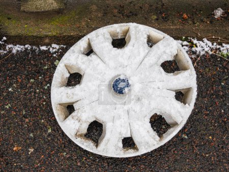An aged car rim rests on the ground, partially covered in a light dusting of snow, reflecting the cold, winter weather in Gothenburg, Sweden. The rim lies abandoned against a backdrop of gravel and sparse winter vegetation, hinting at a story of negl