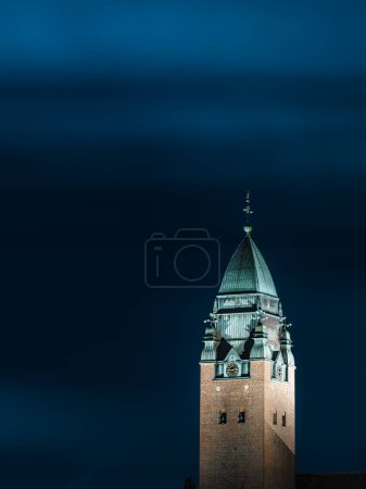 The distinct tower of Masthuggskyrkan stands tall against the darkening evening sky, the intricate architectural details illuminated by strategic lighting, creating a stark contrast with the deep blue backdrop in Gothenburg, Sweden.