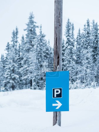 An einem Holzpfahl ist ein blaues Parkschild mit einem weißen P und einem Pfeil angebracht, das die Fahrer zu einem Parkplatz inmitten einer schneebedeckten schwedischen Landschaft führt. Die umliegenden Nadelbäume sind schneegefroren, was die ruhige, winterliche Atmosphäre unterstreicht.