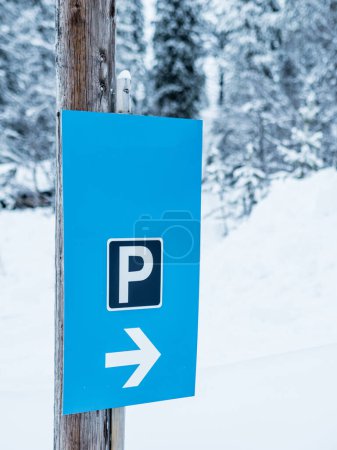 A blue parking sign with a white P and an arrow is affixed to a wooden post, guiding drivers to a parking area amidst a snow-laden Swedish landscape. The surrounding coniferous trees are frosted with snow, highlighting the serene, wintery atmosphere.