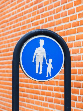 Un letrero peatonal con una silueta de un adulto cogido de la mano con un niño está montado contra una vibrante pared de ladrillo. Destaca el letrero circular azul brillante, que indica un área segura para los peatones, posiblemente cerca de una escuela o zona familiar..