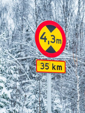 Ein an einer Metallstange montiertes Straßenschild steht an einem Wintertag inmitten einer Schneedecke. Das Schild weist auf eine eingeschränkte Fahrzeughöhe hin und warnt Autofahrer vor möglichen Behinderungen. Der weiße Schnee kontrastiert mit dem rot-weißen Zeichen und schafft eine
