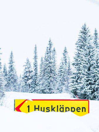 A bright yellow road sign with the inscription 1 Husklappen is displayed prominently against a backdrop of dense, snow-laden coniferous trees in the wintery Swedish landscape, highlighting a location or direction during the seasons snowy embrace.