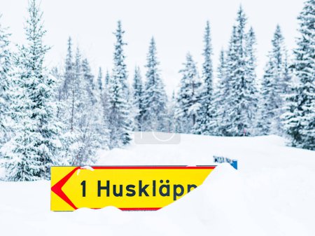 A bright yellow road sign with the inscription 1 Huskalppen is displayed prominently against a backdrop of dense, snow-laden coniferous trees in the wintery Swedish landscape, highlighting a location or direction during the seasons snowy embrace.