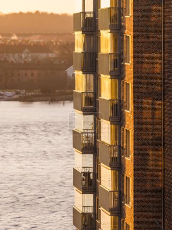 The side of a residential building in Gothenburg glows with the warm light of the setting sun, highlighting its balconies and brickwork. The calm river nearby enhances the tranquil urban scene.