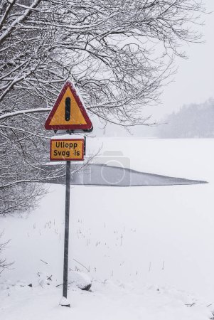 A cautionary triangular sign with an exclamation mark stands out against a backdrop of white, warning of weak ice on a frozen body of water in Sweden during the winter season. Snow-covered trees and ground complete the chilly scene.