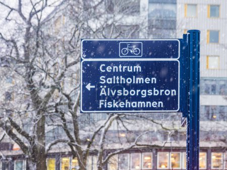 Inmitten des leichten Schneefalls in Göteborg steht ein Fahrradwegweiser, der Radfahrer in Richtung Centrum, Salltholmen, Alvsborgsbron und Fiskehamnen lenkt. Die umgebende Unschärfe der Schneeflocken verleiht der urbanen Umgebung eine heitere Winteratmosphäre.