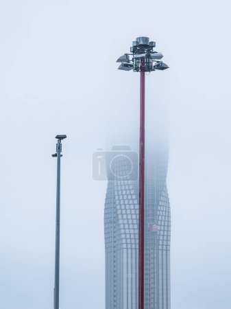 A serene Gothenburg morning finds street lamps standing guard as a towering skyscraper fades into the mist, with the fog lending an ethereal quality to the citys architecture.