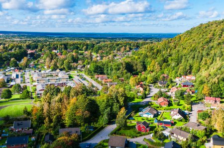An aerial perspective of a town nestled amidst a dense array of trees, showcasing the autumn colors of Sweden. The town is prominently visible against the backdrop of lush green foliage and vibrant fall hues.