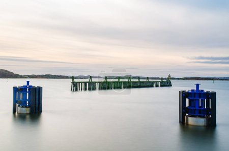 A tranquil scene captures a breakwater in Sweden during dusk. The calm water gently surrounds the wooden structure, displaying a blend of natural and manmade harmony. The muted colors of the sky suggest the peaceful transition from evening to night.