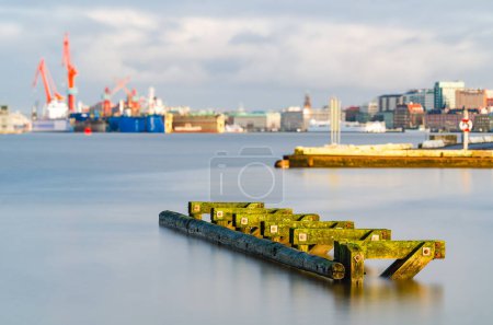 Una serena escena matutina en el puerto de Gotemburgo, donde un muelle de madera cubierto de musgo se extiende en el agua tranquila, con el telón de fondo industrial de las ciudades suavemente borrosa en la distancia bajo un cielo despejado.
