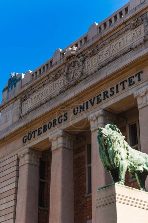 A bronze lion statue stands proudly at the entrance of Gothenburg University in Sweden, symbolizing strength and guardianship. The universitys name is etched in stone above the grand facade, under a clear blue sky, creating an imposing and scholarly 