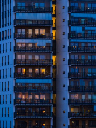A tall building with numerous balconies is illuminated at night, showcasing the glowing lights from within the rooms and on the balconies. The architecture stands out against the dark sky, creating a striking urban scene.