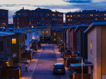 The image shows a street at dusk, flanked by identical row houses. The sky is a deep blue hue, with streetlights beginning to illuminate the scene.