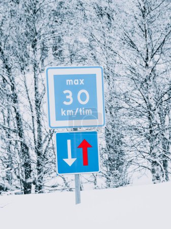 A blue and white speed limit sign displays a maximum of 30 kilometers per hour along a road and priority must be given to vehicles in opposite direction. The signs, adorned with a snow layer, is set against a backdrop of densely snow-laden trees, hig