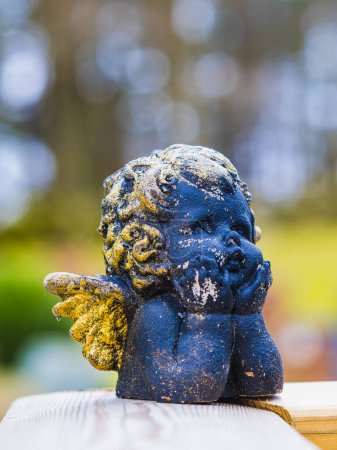 Una estatua de ángel azul envejecido con alas doradas. El fondo presenta verdes y amarillos suavemente borrosos, lo que sugiere un entorno de jardín bañado por la luz natural de un día soleado..