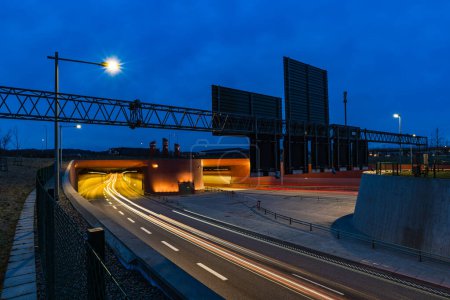 Alors que le crépuscule s'installe à Gothenburg, en Suède, des véhicules sont capturés en mouvement entrant et sortant d'un tunnel très éclairé. Le contraste entre l'éclairage chaud du tunnel et le bleu frais du ciel nocturne met en évidence l'infrastructure urbaine.