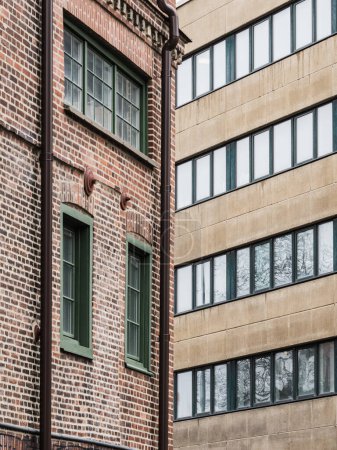 Ein hoher Backsteinbau mit grünen Fenstern steht neben einem schlanken, modernen Gebäude. Der Kontrast in der Architektur schafft eine dynamische Stadtlandschaft.