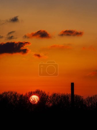 Foto de The sun sets behind the trees of a forest in Gothenburg, Sweden, casting a warm orange glow over the scene. A single smokestack stands tall above the treetops, silhouetted against the colorful sky. - Imagen libre de derechos