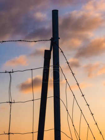 A silhouette of a barbed wire fence stands against a vibrant sunset sky with soft clouds in Gothenburg, Sweden. The fence post anchors the wire in a peaceful yet poignant testament to rural boundaries at dusk.