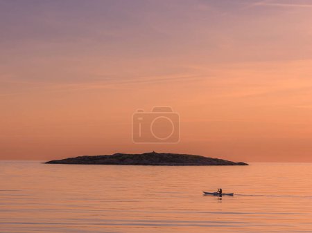 A woman is seen gracefully kayaking across the smooth, reflective surface of the sea during a tranquil sunset in Sweden. The golden hues of the setting sun create a serene backdrop, silhouetting the solo kayaker in a peaceful maritime scene.