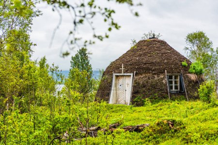 A traditional hut, covered in natural materials, stands amidst lush greenery in Lappland, Sweden. The untouched landscape and cloudy sky contribute to the wild, serene atmosphere.