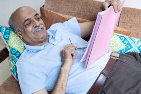Photo for Indian senior man Reading book while lay down at sofa at living room,Retirement Life - Royalty Free Image