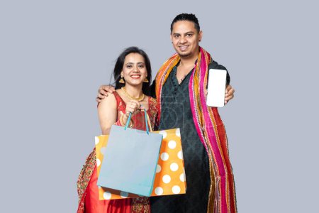 Photo for Indian Young couple in ethnic wear standing against grey background holding Shopping bags showing and pointing at empty phone screen. - Royalty Free Image