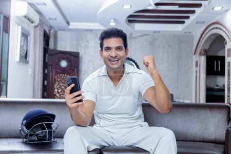 Téléchargez les photos : Homme heureux en robe de cricket célébrant victoire assis sur le canapé au salon regardant vers le téléphone mobile, Homme appréciant regarder le cricket à la maison - en image libre de droit