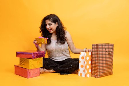 Photo for Portrait of Excited Asian indian woman sitting on ground with shopping bags & Giftbox isolated on orange background, Shopper or shopaholic concept - Royalty Free Image
