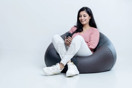 Photo for Indian latin female siting on bean bag and watching video on mobile , copy space - Royalty Free Image