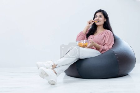 Photo for Happy Beautiful Female sitting on Bean bag and having chips snacks watching Movies on Isolated background - Royalty Free Image